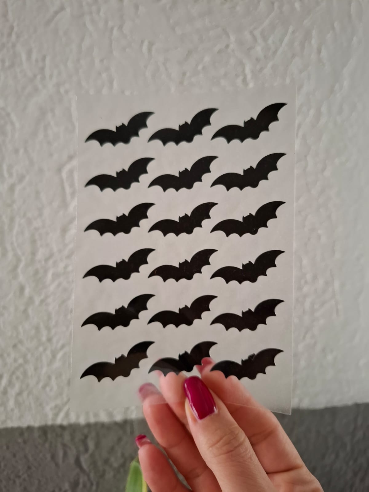 Eine Hand mit roten Nägeln hält ein transparentes Blatt, das mit zahlreichen schwarzen Fledermausaufklebern verziert ist und so vor der strukturierten weiß-grauen Wand für eine gruselige Halloween-Stimmung sorgt.
A hand with red nails holds a transparent sheet adorned with numerous black bat stickers, creating a spooky Halloween vibe against the textured white and grey wall.